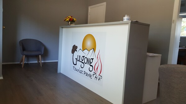 A reception desk with a logo for 'Julgong Tourist Park'. There is a chair to the left and a small table with a vase of flowers on top of the desk.
