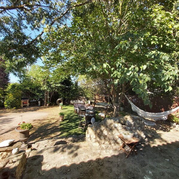 Un área de estar al aire libre con mesas y sillas bajo un árbol, rodeada de follaje.