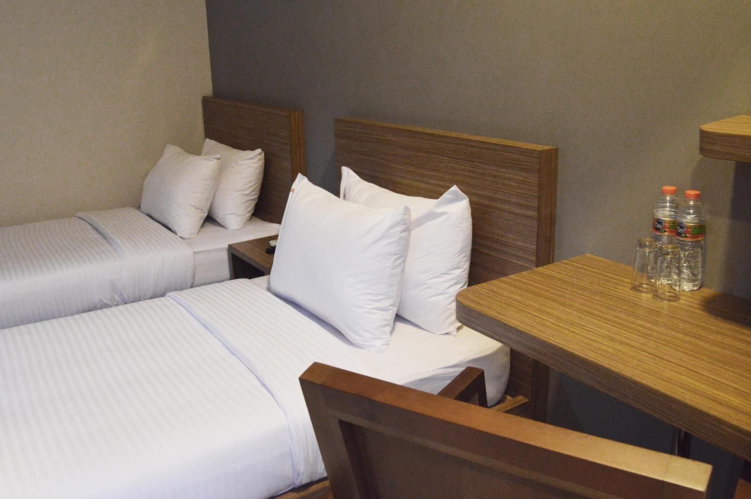 Une chambre d’hôtel avec deux lits simples, chacun agrémenté de draps et d’oreillers blancs, une table de chevet en bois avec une bouteille et des verres, et un bureau en bois également avec une bouteille et des verres.