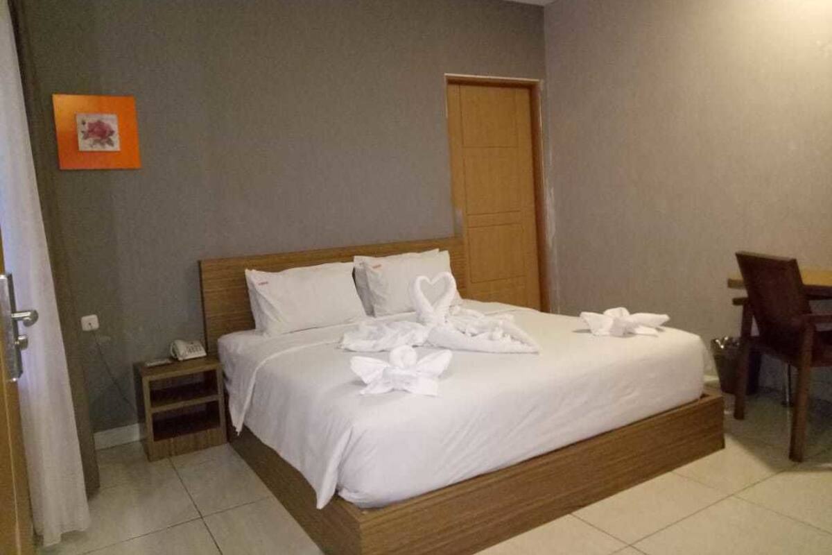 Una cama bien hecha con sábanas blancas y adornos de cisnes, una mesita de noche de madera con un teléfono, una puerta de madera y una silla en una habitación de hotel.