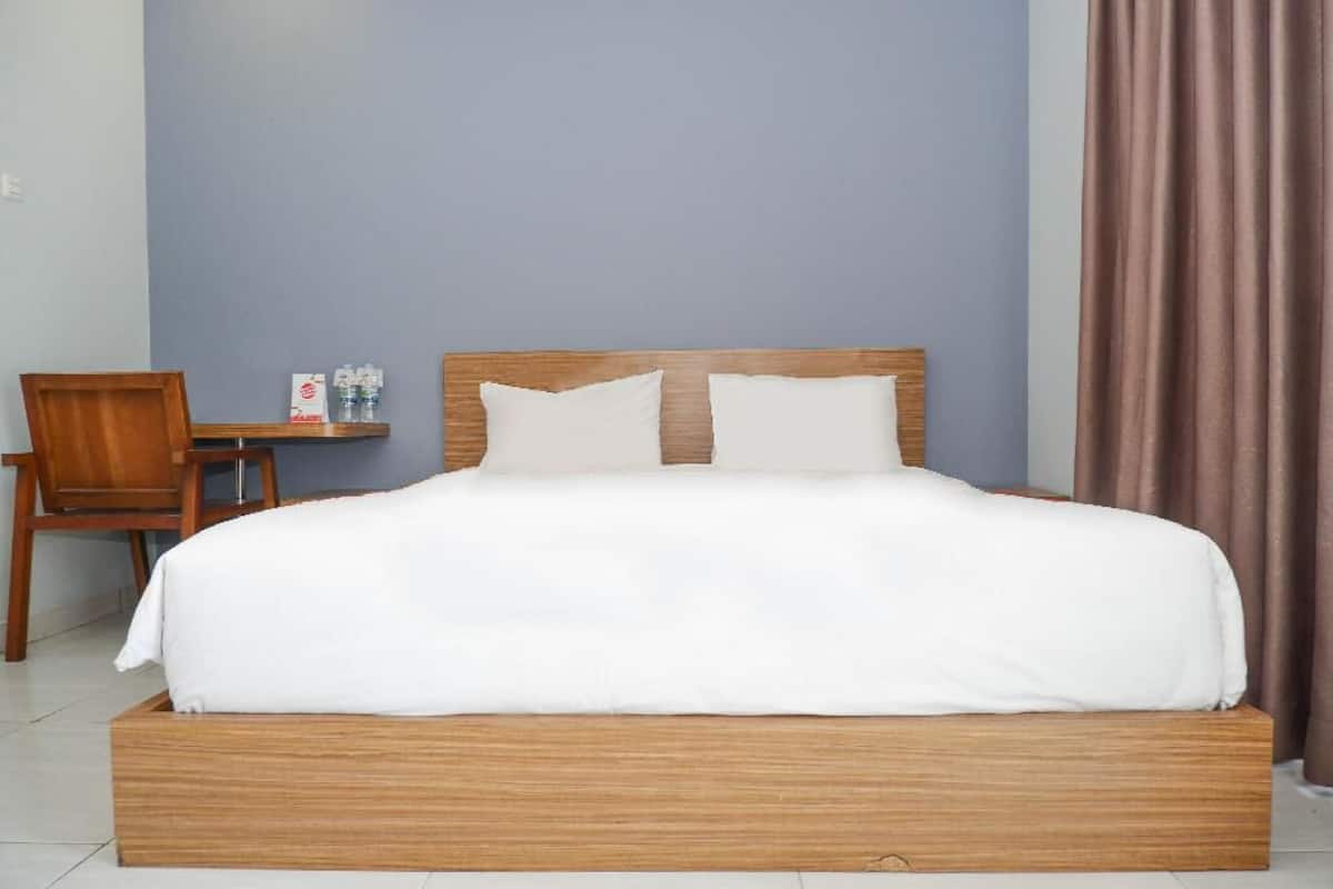 Una cama bien hecha con estructura de madera y ropa de cama blanca, una silla de madera y una mesita con botellas de agua y una caja.
