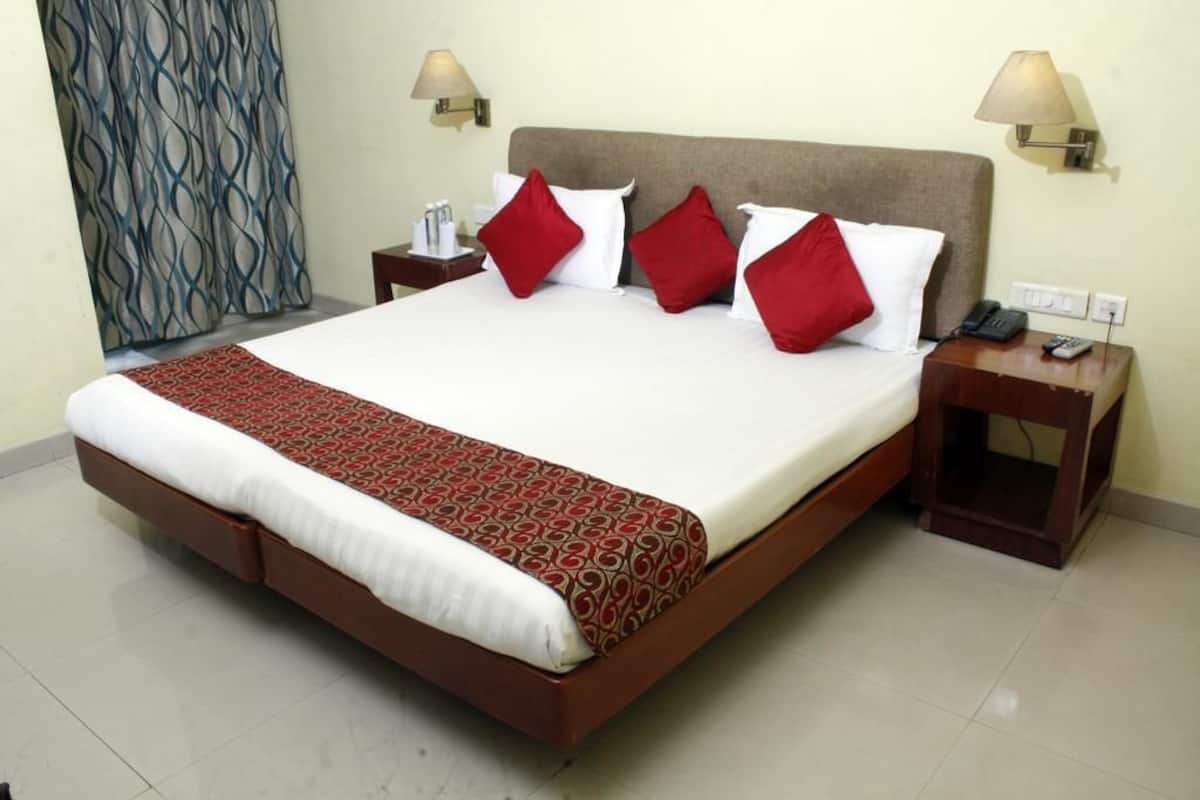Una habitación de hotel con una cama grande, almohadas rojas y blancas, una colcha estampada, dos mesitas de noche con lámparas y una cortina de estampado ondulado.