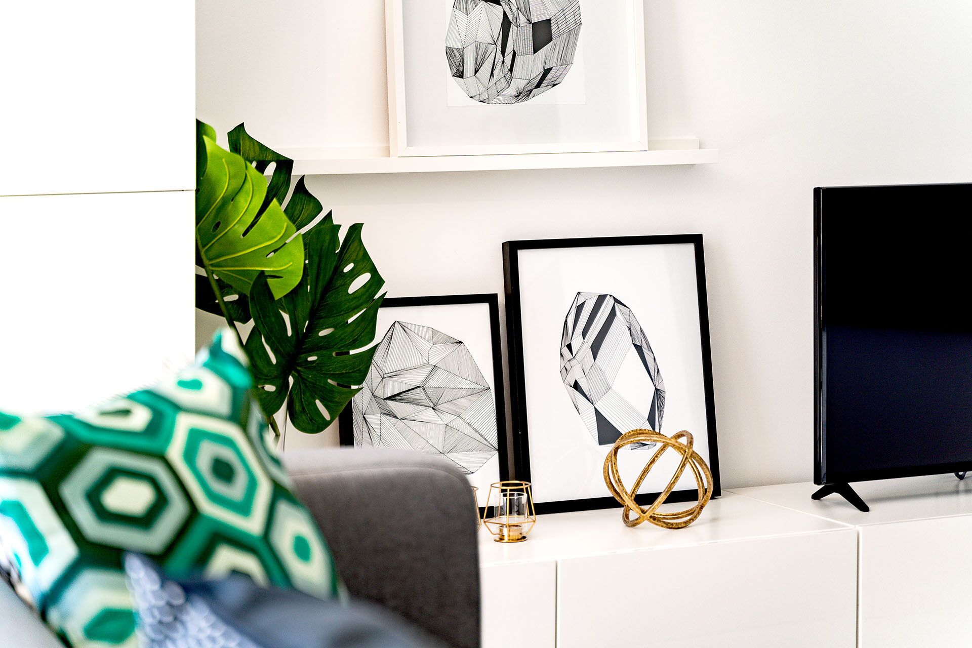 Ein modernes Wohnzimmer mit einer Couch, einem Couchtisch und einem Fernsehschrank. An den Wänden hängen gerahmte Kunstwerke und in der Ecke steht eine große grüne Pflanze.