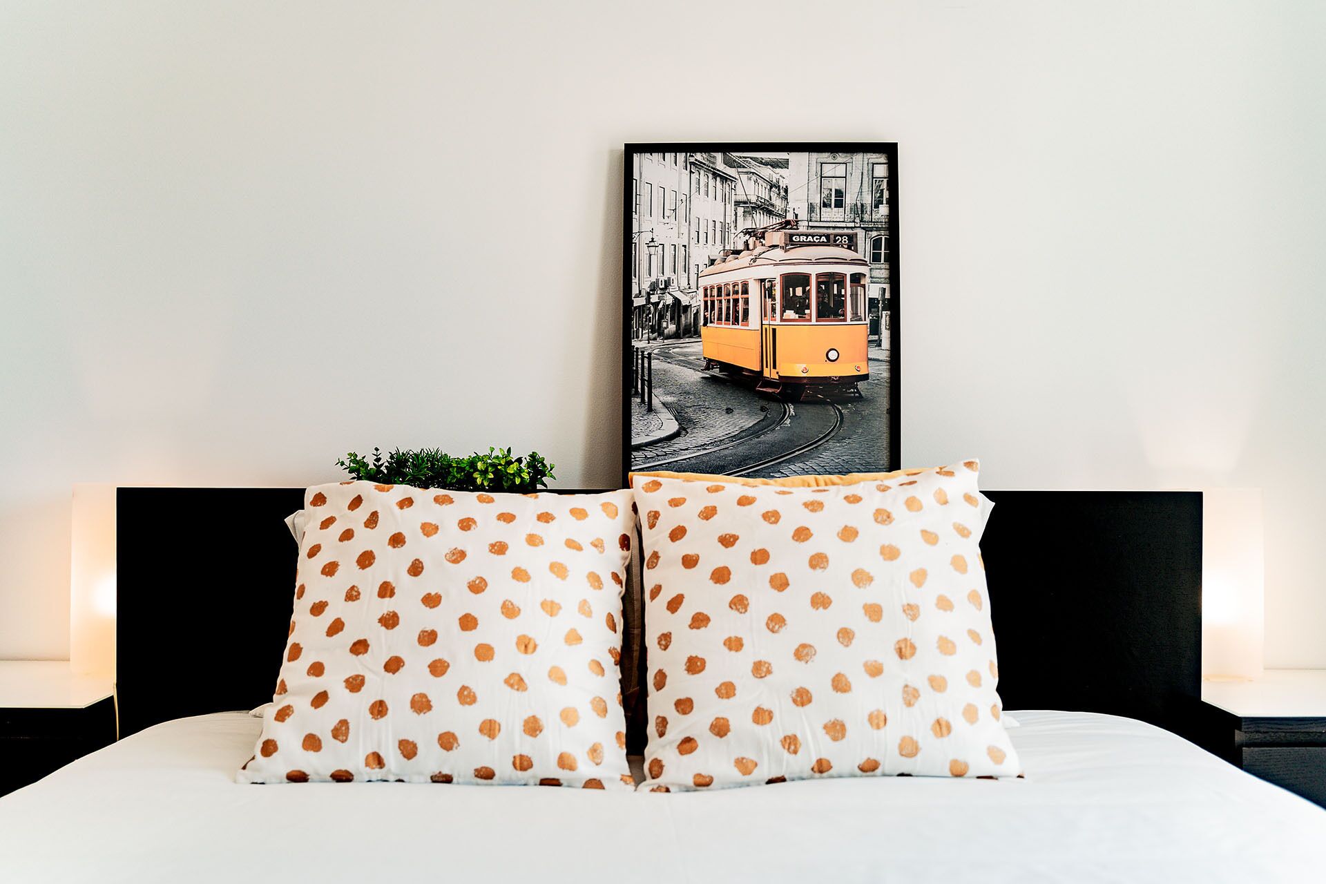 A bed with white bedding and two pillows with a polka dot pattern. A framed picture of a tram is mounted above the bed.