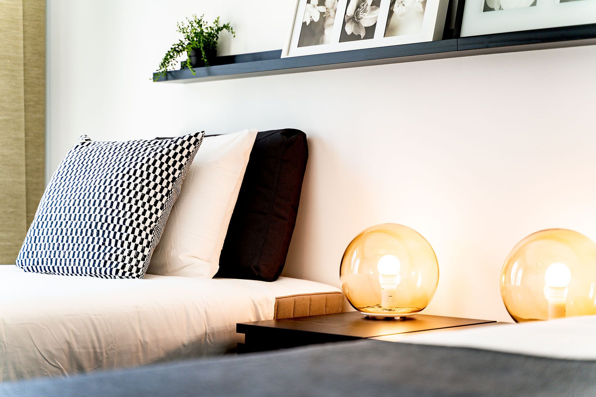 Un dormitorio ordenado con una cama, una mesita de noche con dos lámparas esféricas, un cojín estampado en blanco y negro, y una estantería sobre la cama con una planta pequeña y fotografías enmarcadas.