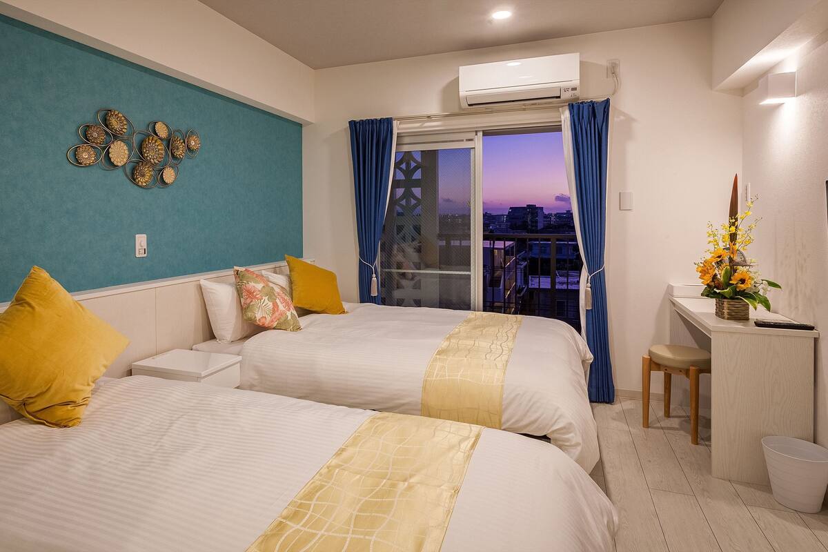 Ein Hotelzimmer mit zwei Betten, einem Schreibtisch mit Stuhl, einem Fenster mit Vorhängen, einer Klimaanlage und einem Wandbild.