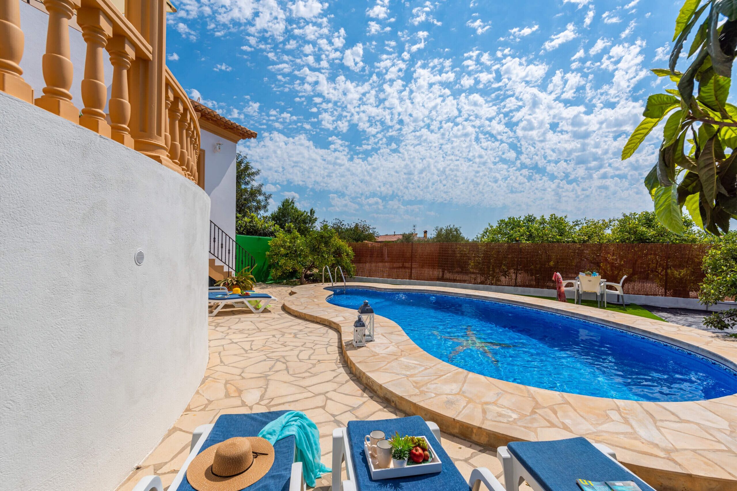 Piscina com ladrilhos de pedra, cadeiras de descanso e mesa com chapéu e bandeja de frutas.