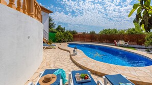 Piscina com ladrilhos de pedra, cadeiras de descanso e mesa com chapéu e bandeja de frutas.