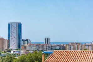 A cityscape with a prominent tall building with a blue facade and a red cross design, surrounded by other residential buildings and a clear sky. - Villa Serena - PlusHolidays (Calpe)