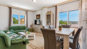 Uma sala de estar moderna com um sofá verde, uma mesa de jantar com cadeiras, uma TV e vista para a cidade através de grandes janelas.