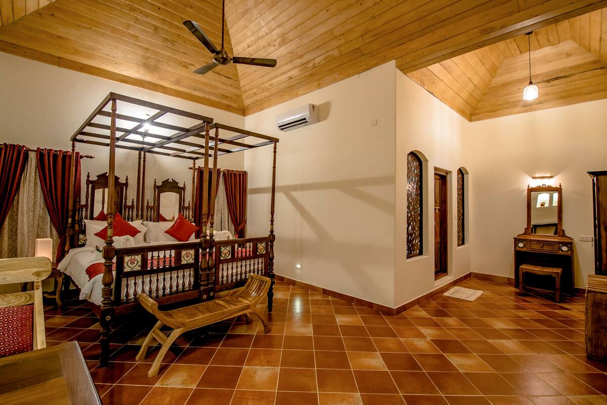 Een hemelbed met houten frame en rode kussens, een houten bankje en een plafondventilator in een kamer met betegelde vloer en houten balken.