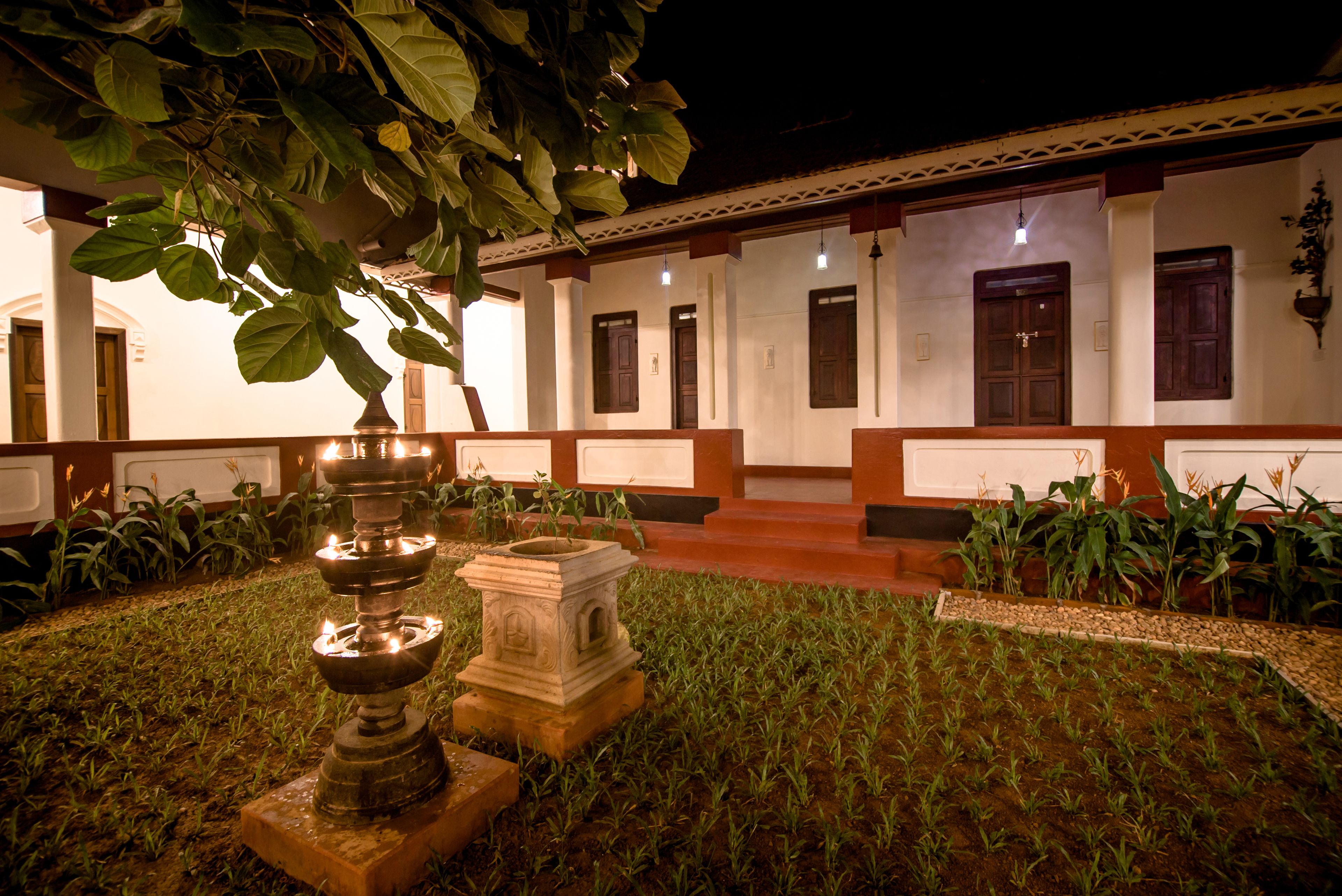 A courtyard with a stone fountain, a well, and a pathway leading to a building with multiple doors and windows.