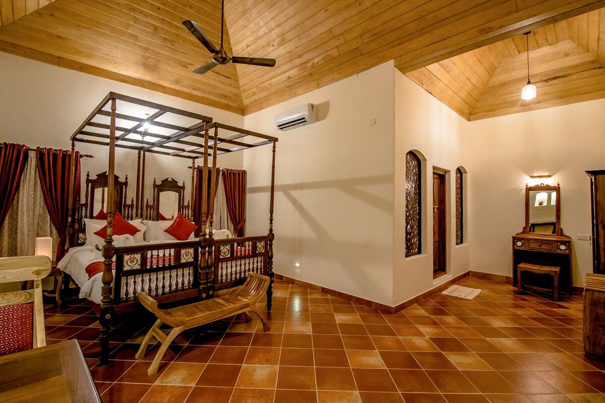 Een hemelbed met houten hemel, een bankje en een klein tafeltje in een kamer met betegelde vloer en houten balkenplafond.