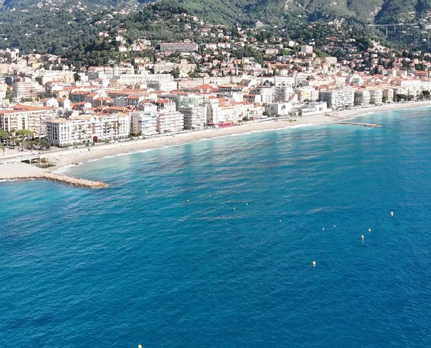 A coastal cityscape with buildings along the waterfront, a clear blue sea, and mountains in the background.