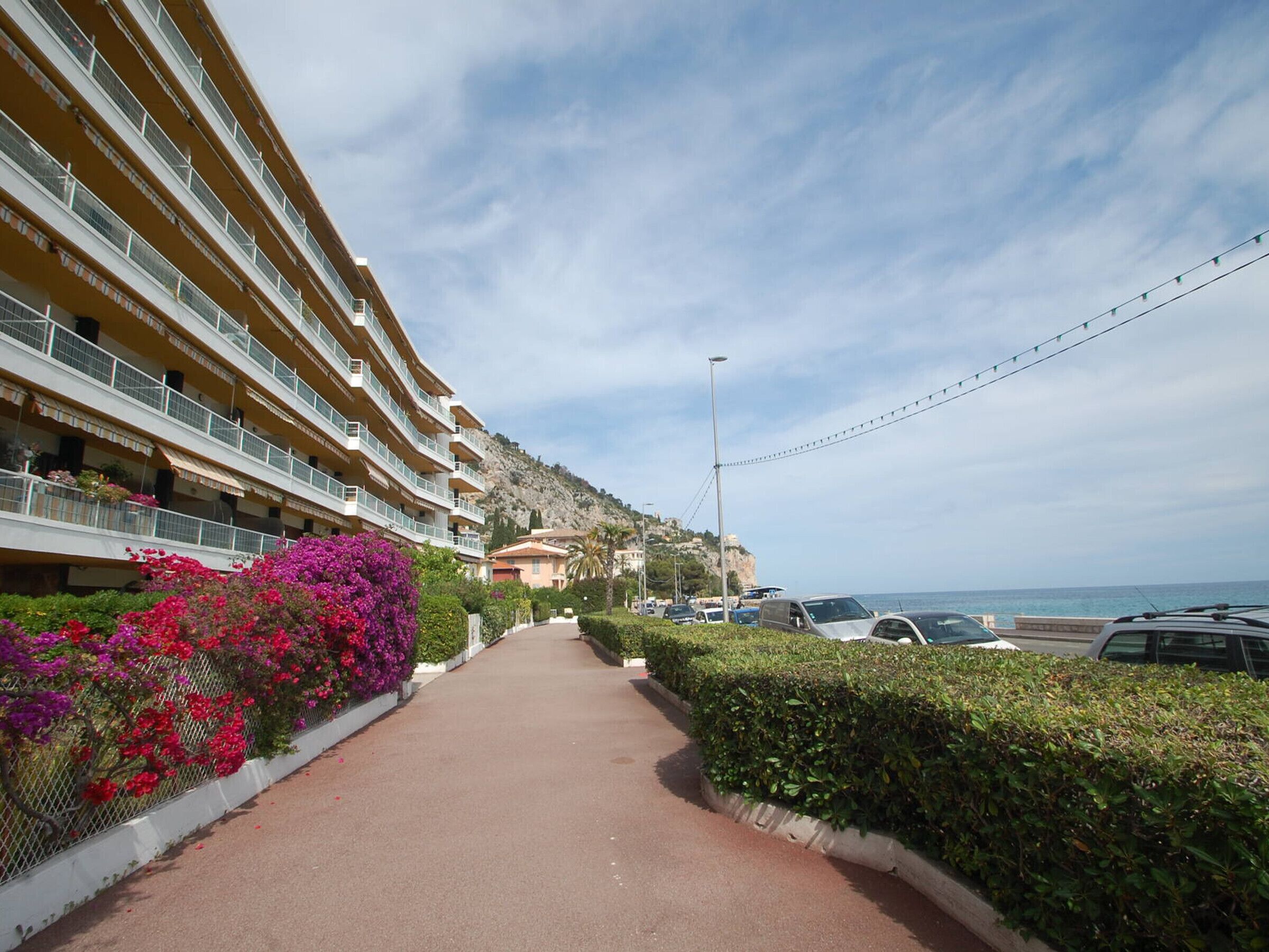 Un camí que condueix a un hotel de diverses plantes amb balcons, envoltat de verdura i vistes al mar.