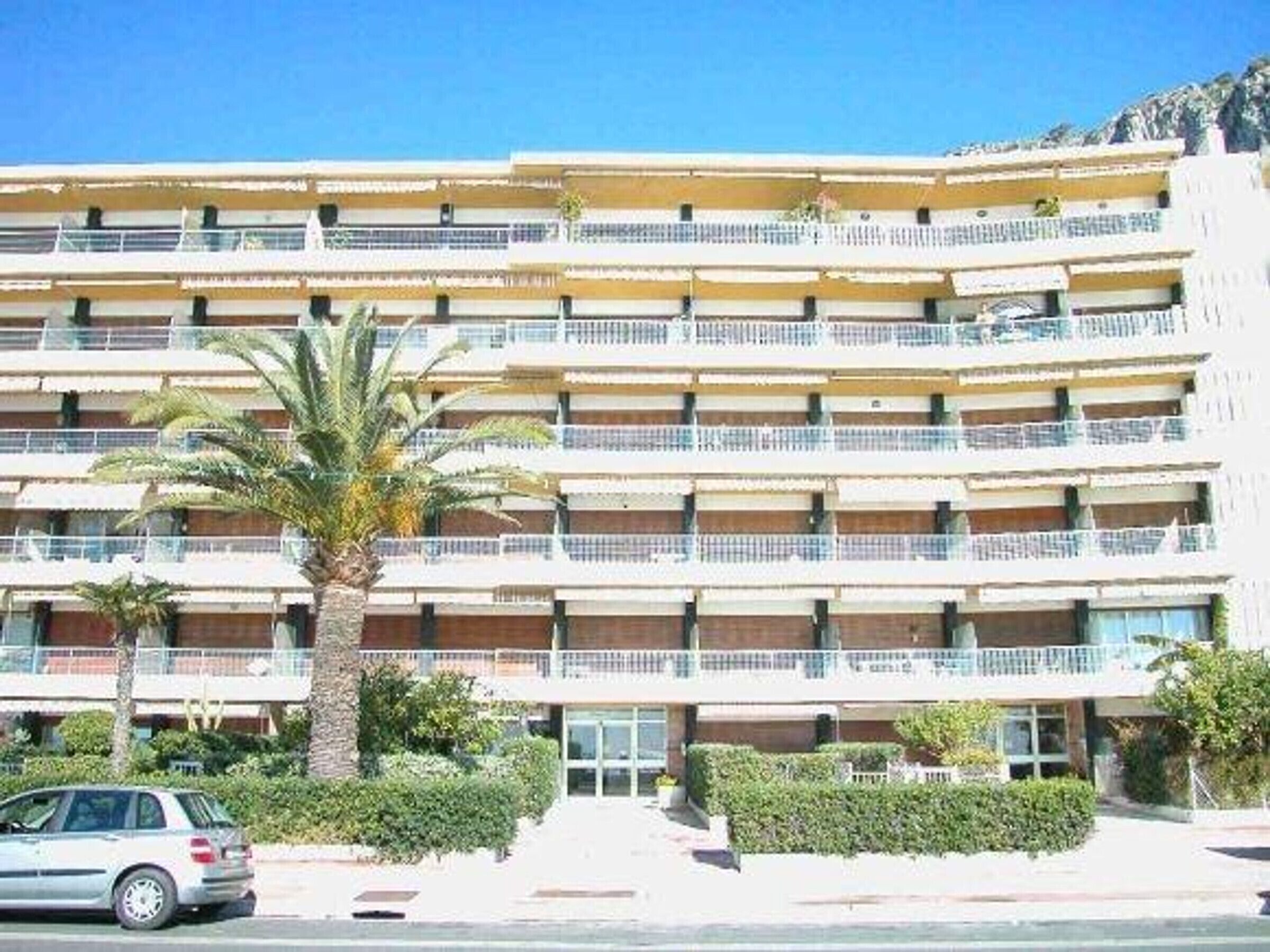 A multi-story hotel with balconies, a car parked in front, and a palm tree.