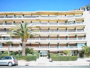 Hotel com vários andares e varandas, um carro estacionado na frente e uma palmeira.