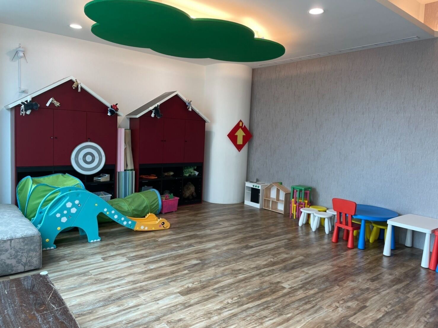 A spacious room with a wooden floor, a play area with a slide and climbing toys, a red playhouse, and a small white table with colorful chairs.