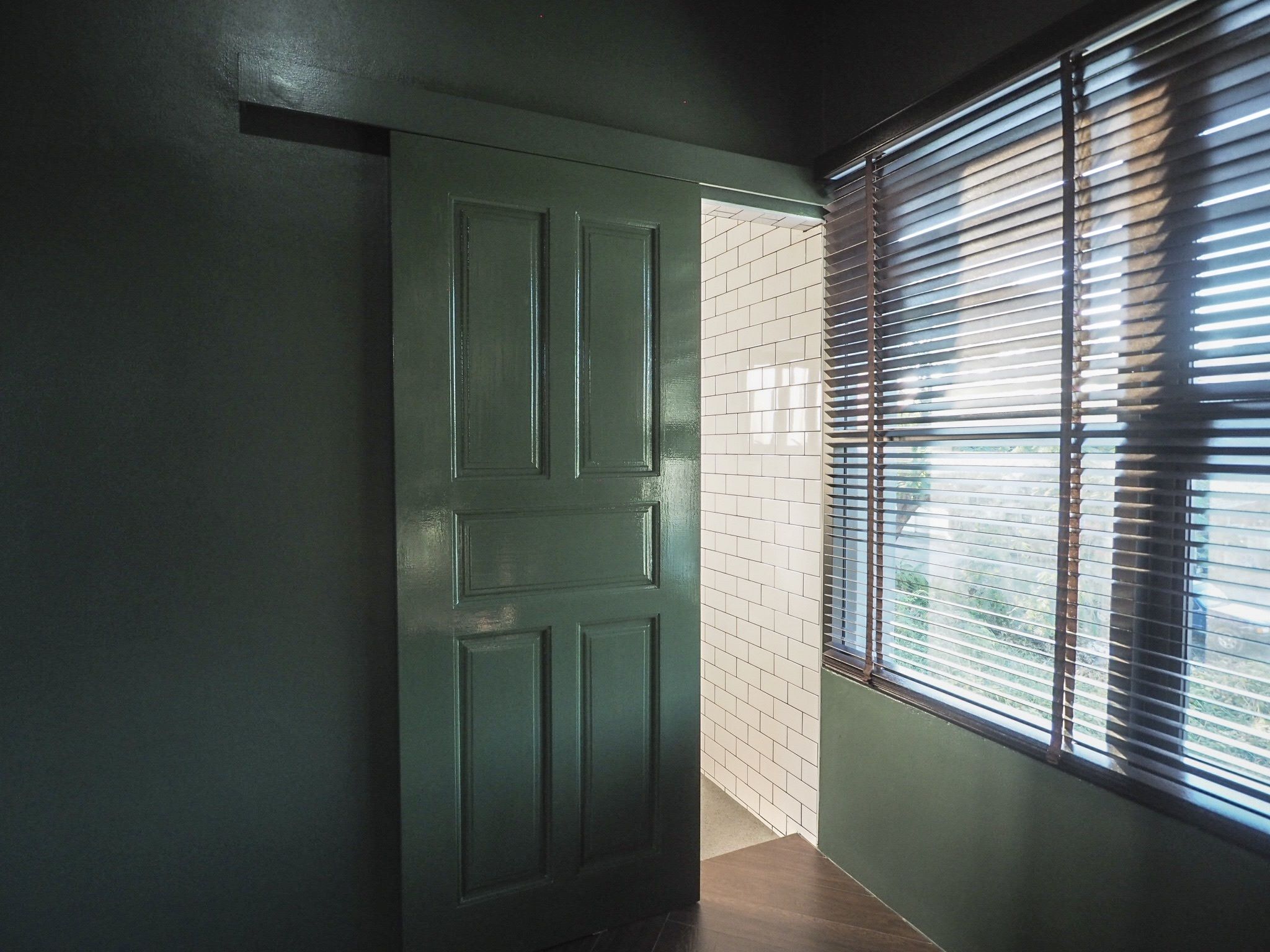 A hallway with a green door, a window with blinds, and a tiled wall.