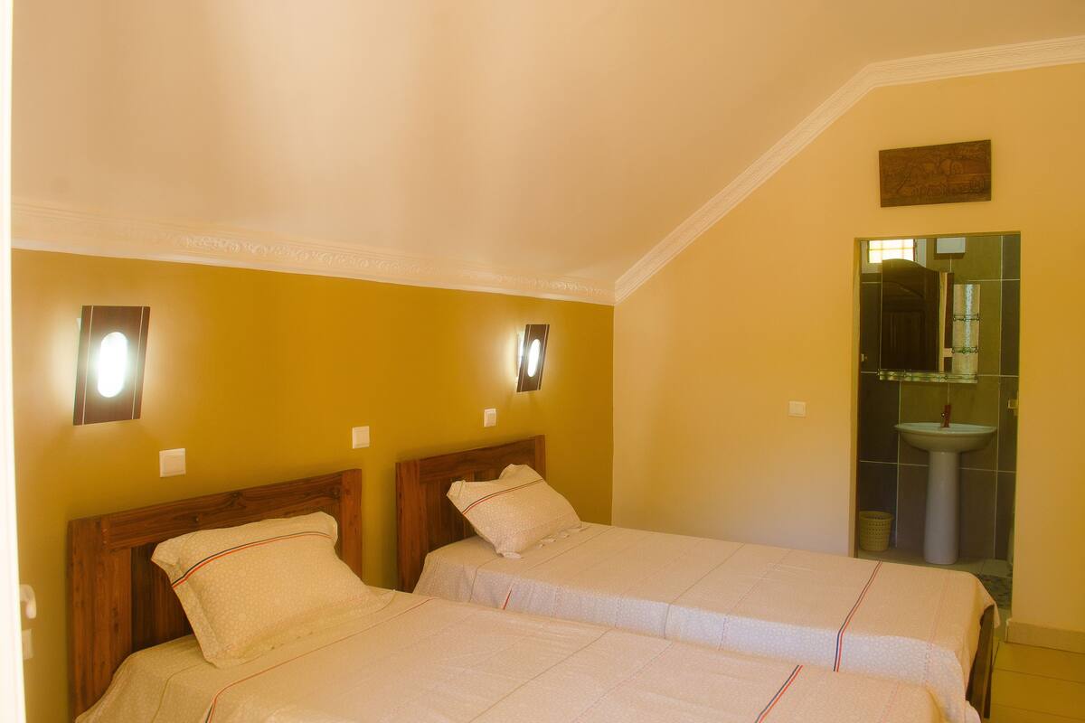 A hotel room with two single beds, a wooden headboard, a wall-mounted light, and a bathroom with a sink and mirror.