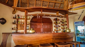 A bar with wooden shelves and stools, a large wooden ship-like structure, and a refrigerator with beverages. - Laguna Beach (Morondava)