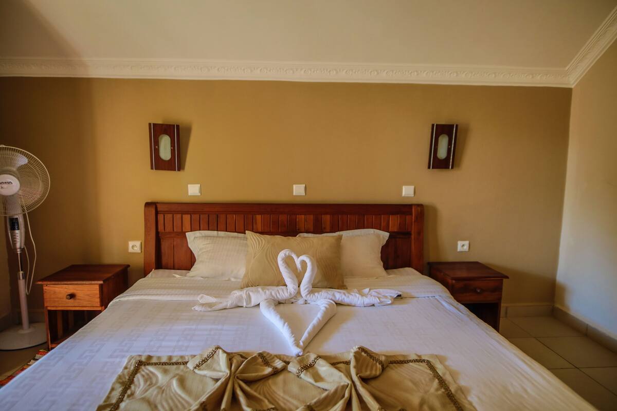 A hotel room with a large bed, wooden headboard, two bedside tables, a standing fan, and two wall-mounted picture frames.