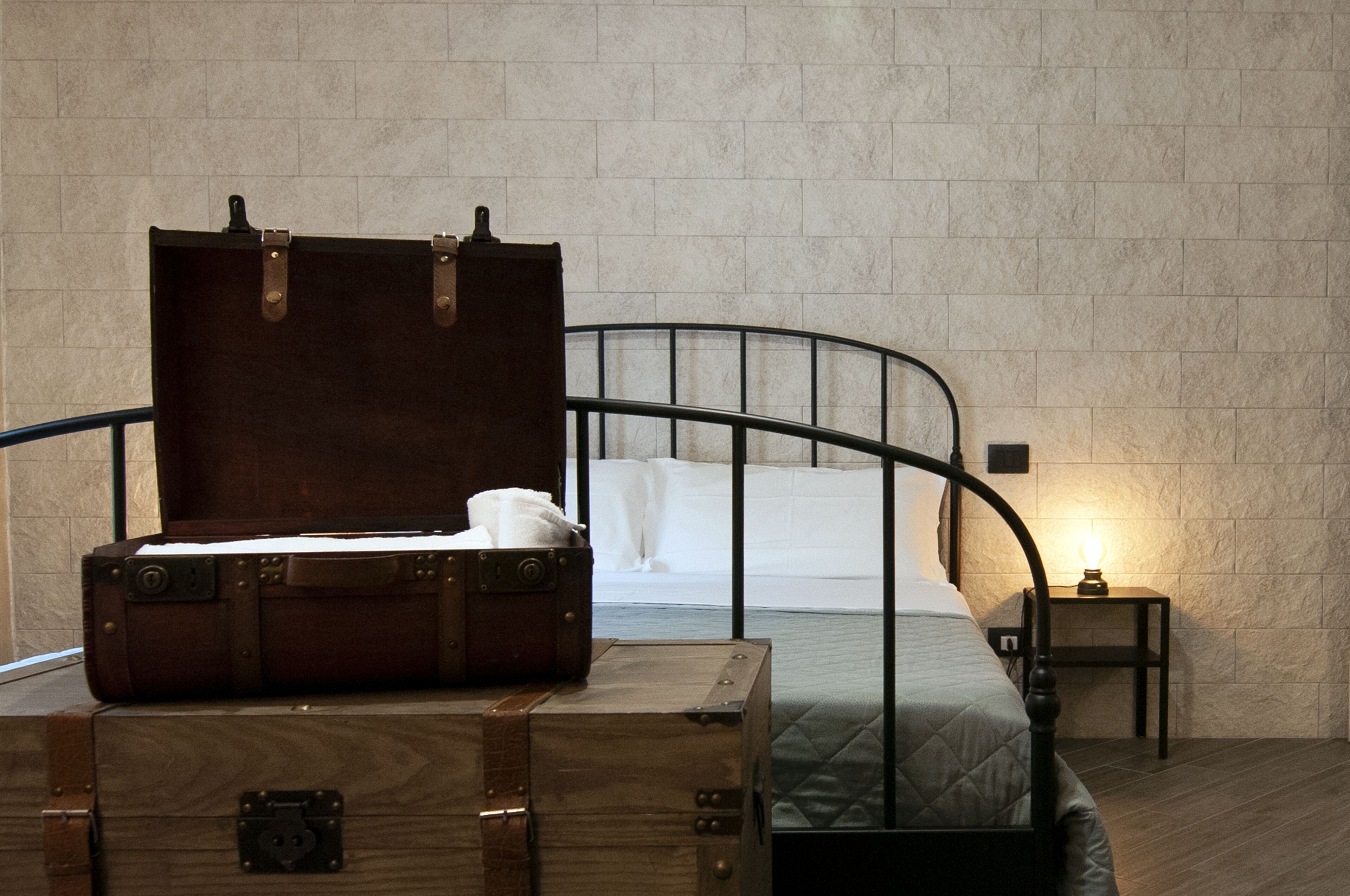 Una habitación con una cama de estructura metálica, una mesita de noche con lámpara y un cofre de madera con una maleta.