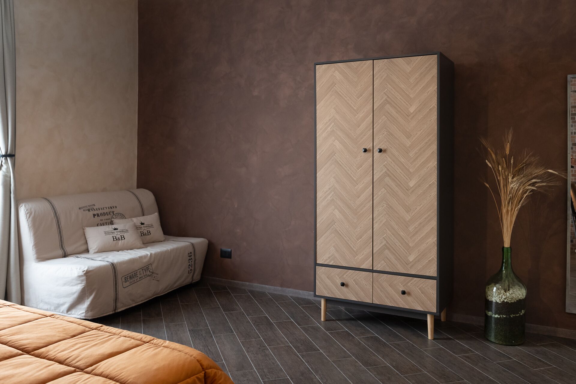 A bedroom with a bed, a sofa, a wardrobe, and a vase with dried plants.