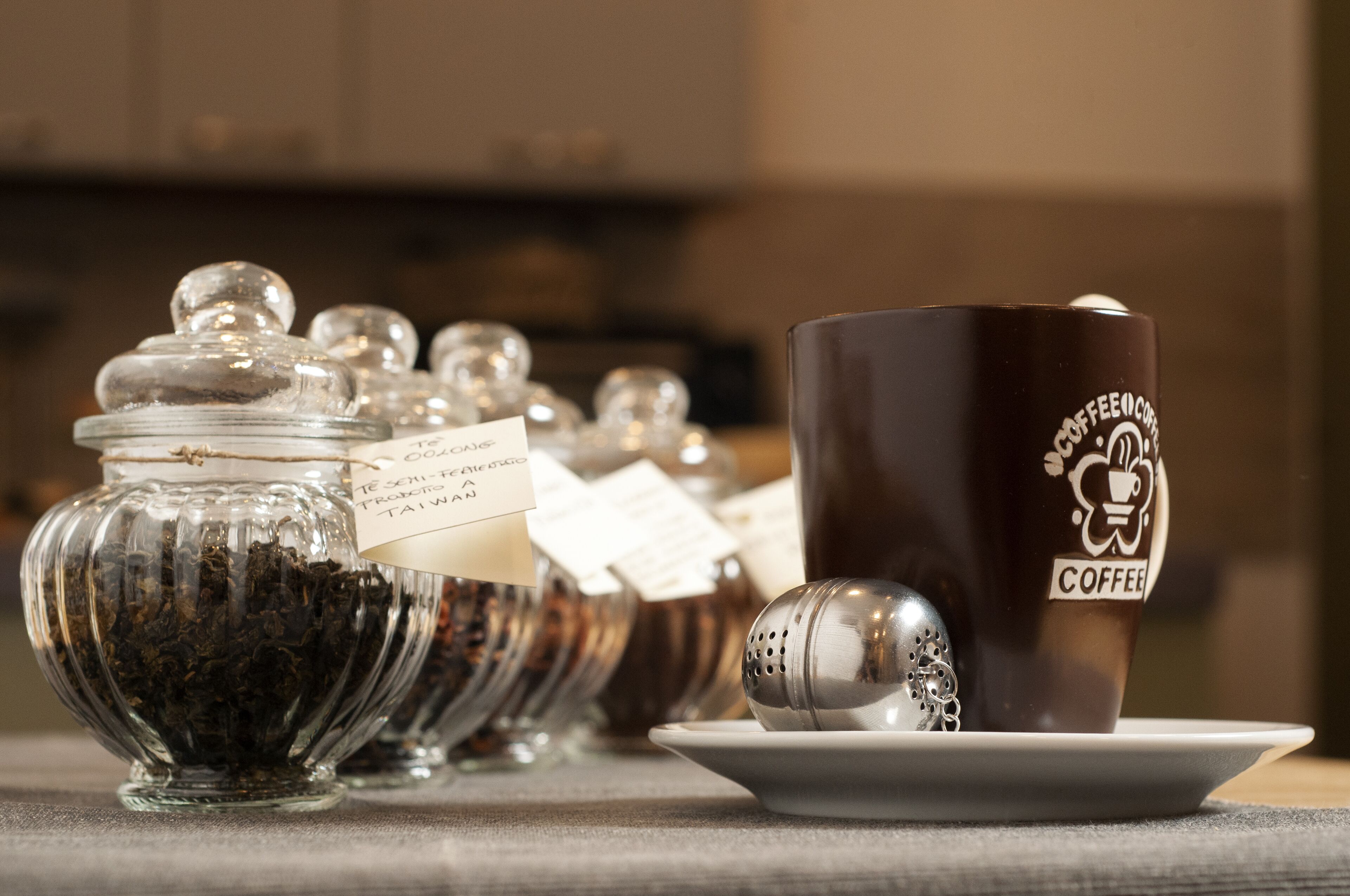 A coffee cup with a logo and a tea ball on a saucer, next to a glass jar with tea leaves and a tag.