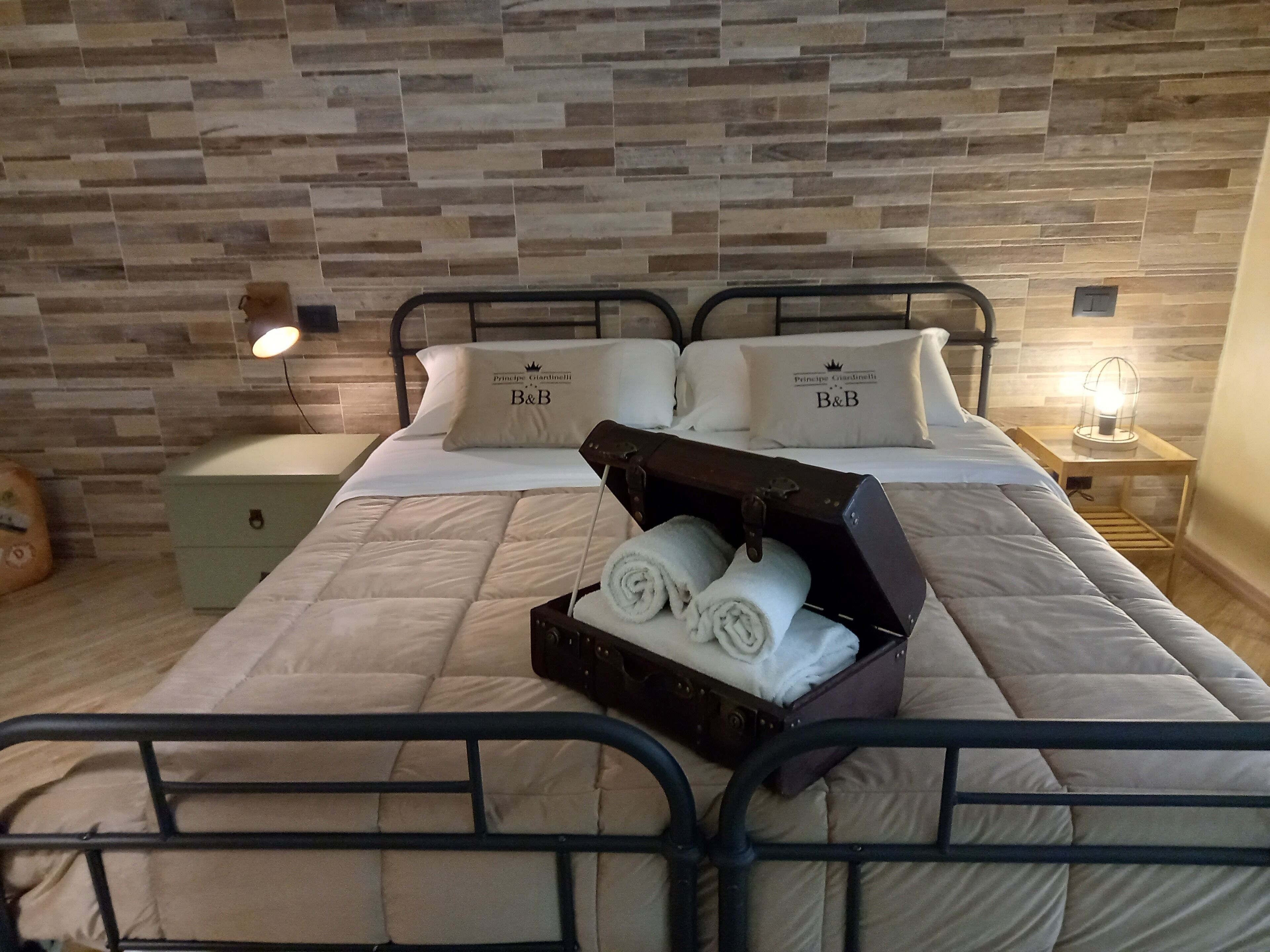 Una cama bien hecha con una colcha acolchada, dos almohadas con el logotipo 'B&B', una maleta con toallas y una mesita de noche con una lámpara.