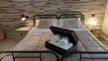 Una cama bien hecha con una colcha acolchada, dos almohadas con el logotipo 'B&B', una maleta con toallas y una mesita de noche con una lámpara.