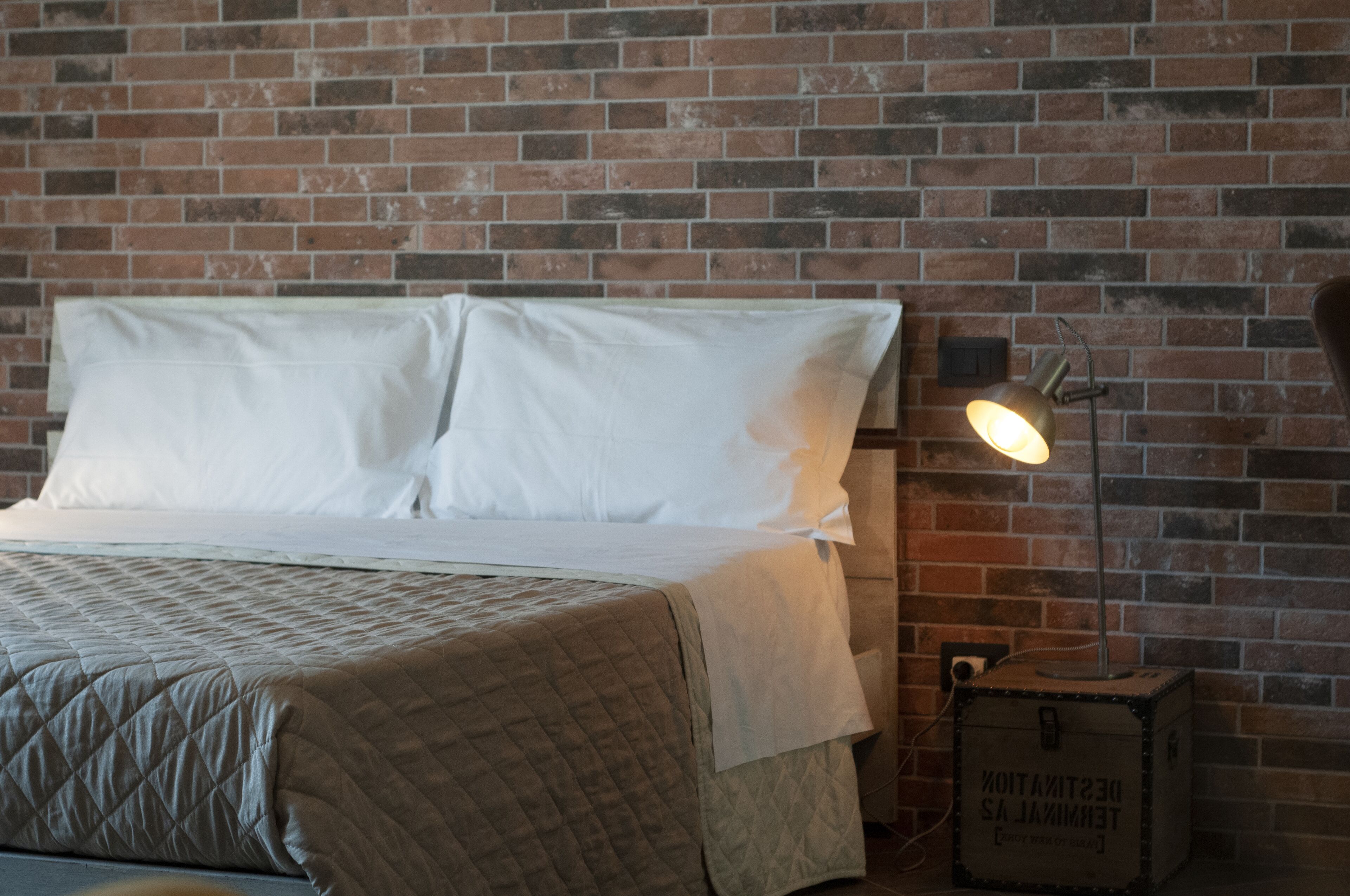 A bed with white linens and a quilted cover, a bedside lamp, and a brick wall.