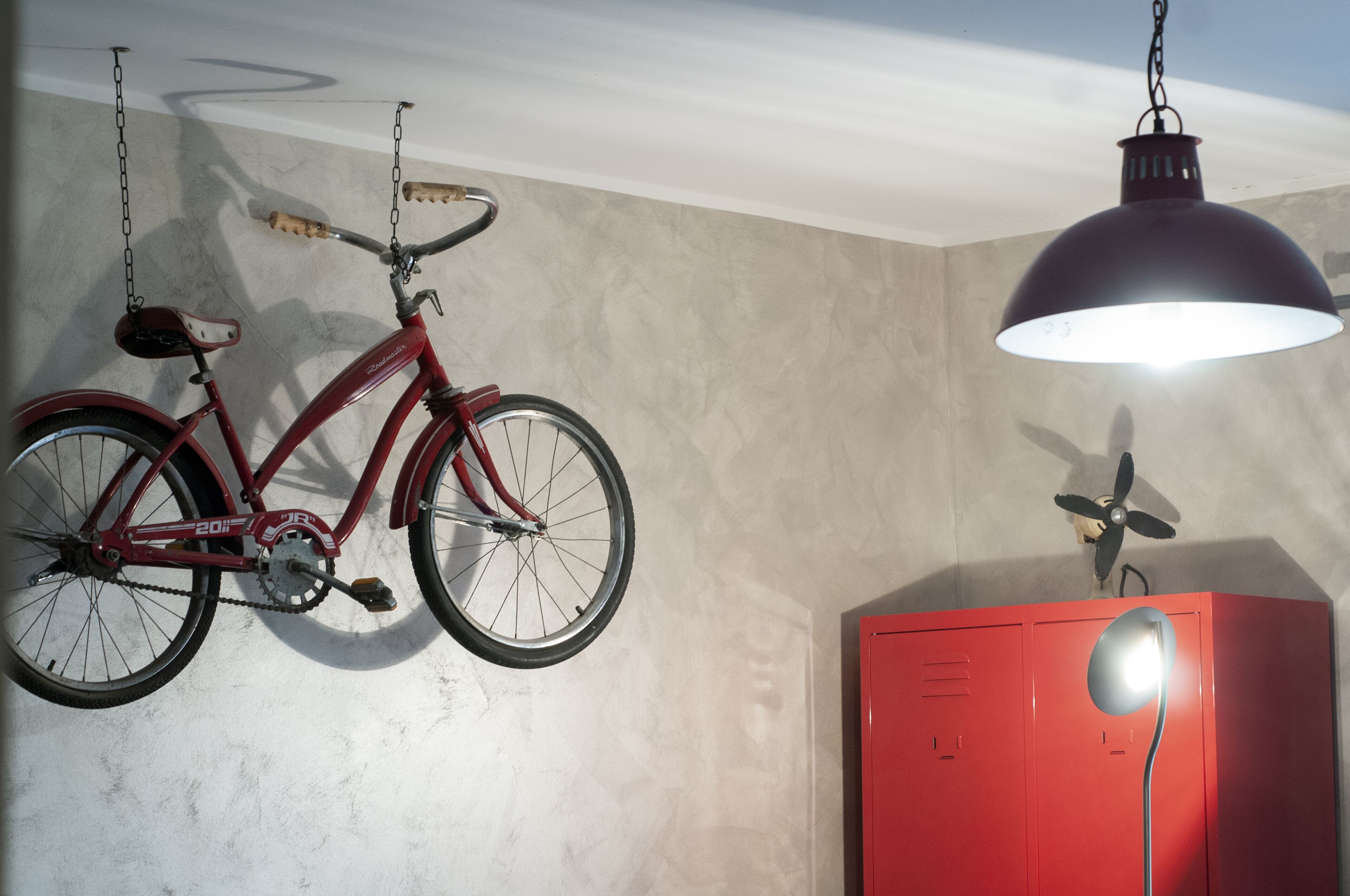 Een rode fiets hangt aan het plafond, een rode kast met een ventilator, en een hanglamp.