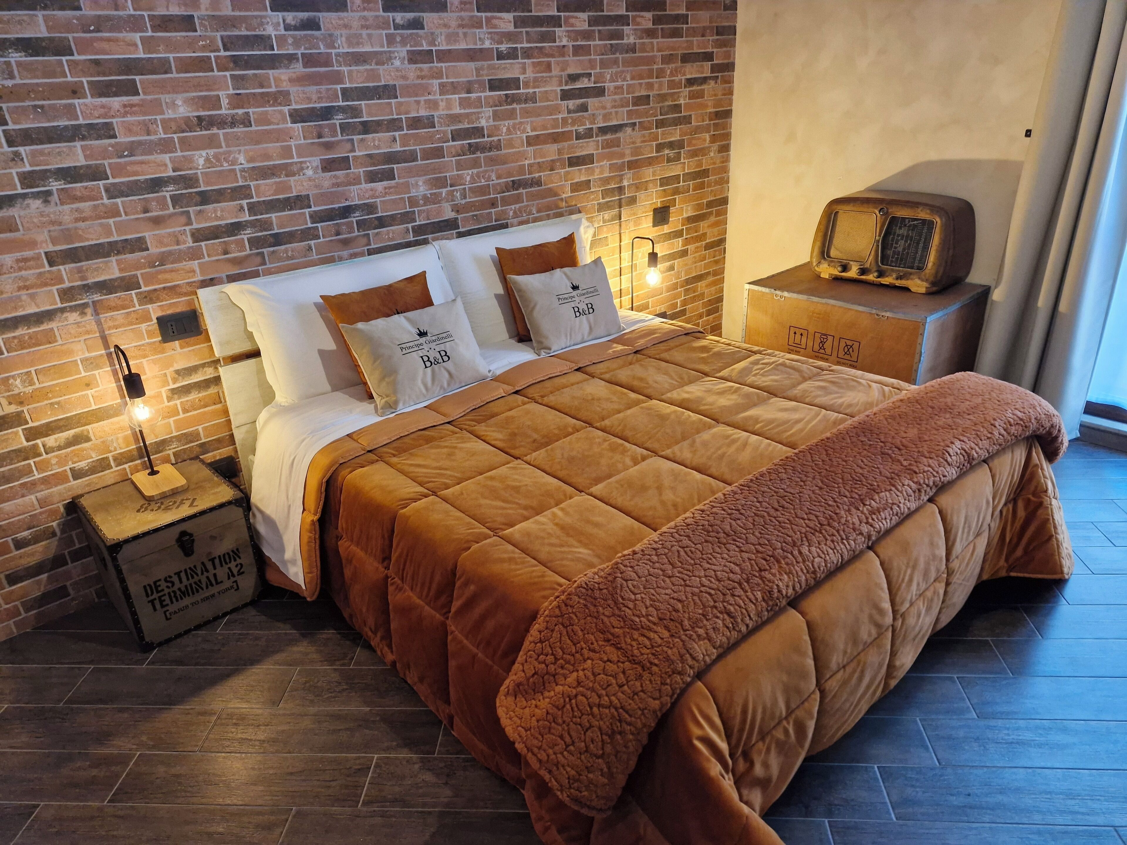 A bedroom with a brick wall, a wooden nightstand, a vintage radio, and a bed with a brown quilt.
