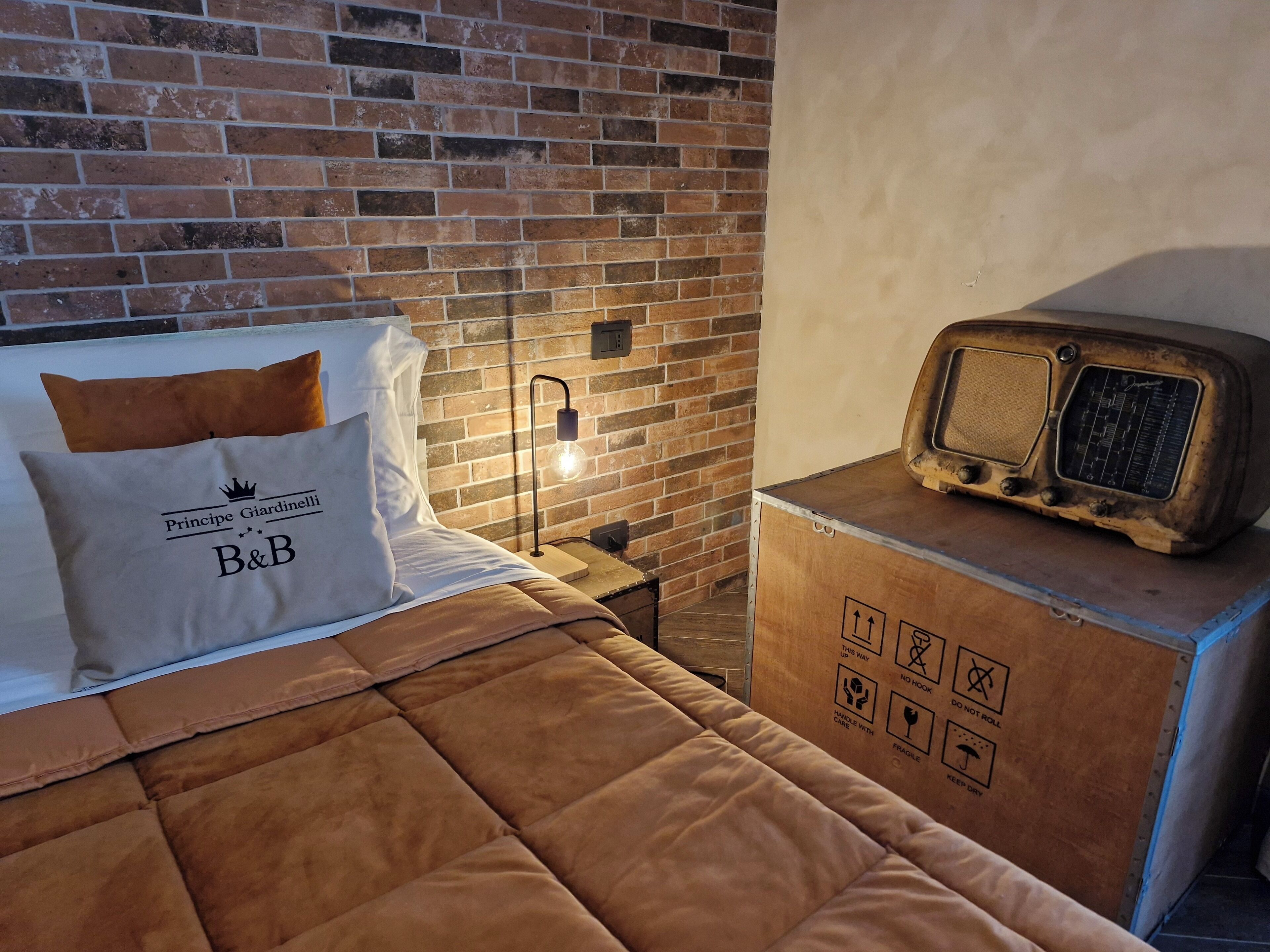 A bed with a brown quilt, a pillow with "Principe Giardinelli B&B" text, a brick wall, a nightstand with a lamp, and a vintage radio.