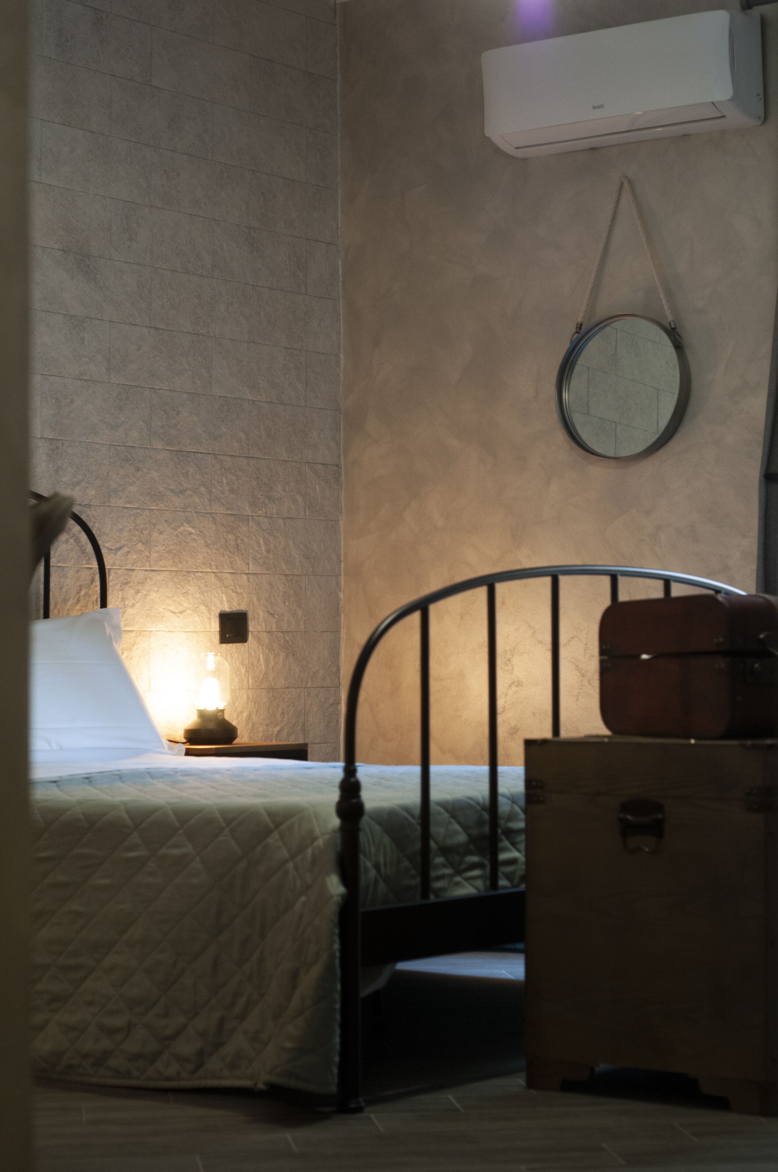 A bedroom with a bed, bedside lamp, wall-mounted air conditioner, and a round mirror.
