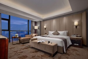 A modern hotel room with a large bed, a sofa, a bench, and a nightstand. There is a city view through large windows.