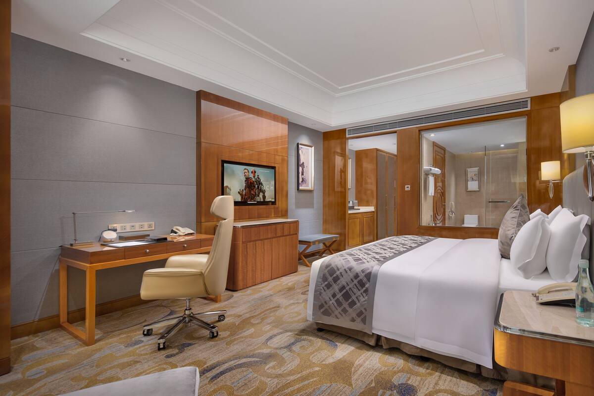 A hotel room with a wooden desk, a comfortable chair, a bed with a patterned bedspread, and a large mirror.