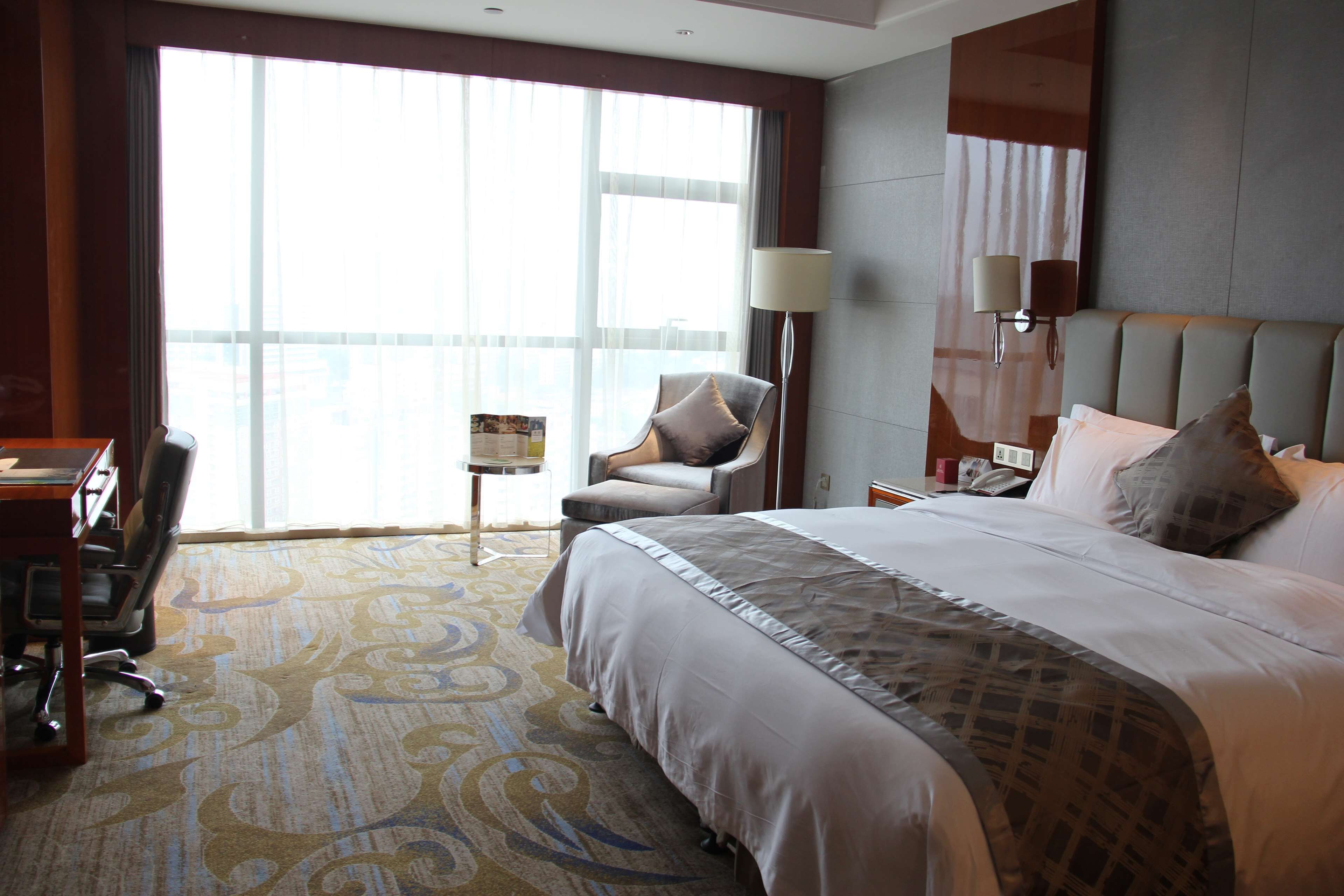 Habitación de hotel con una cama grande, un escritorio, una silla, una lámpara y vistas de la ciudad a través de amplias ventanas.