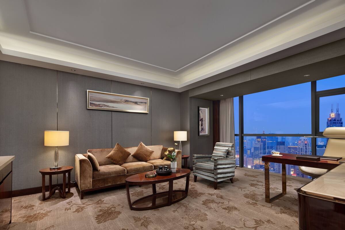 A modern hotel room with a large window offering a city view, a sofa, a chair, and a small table.