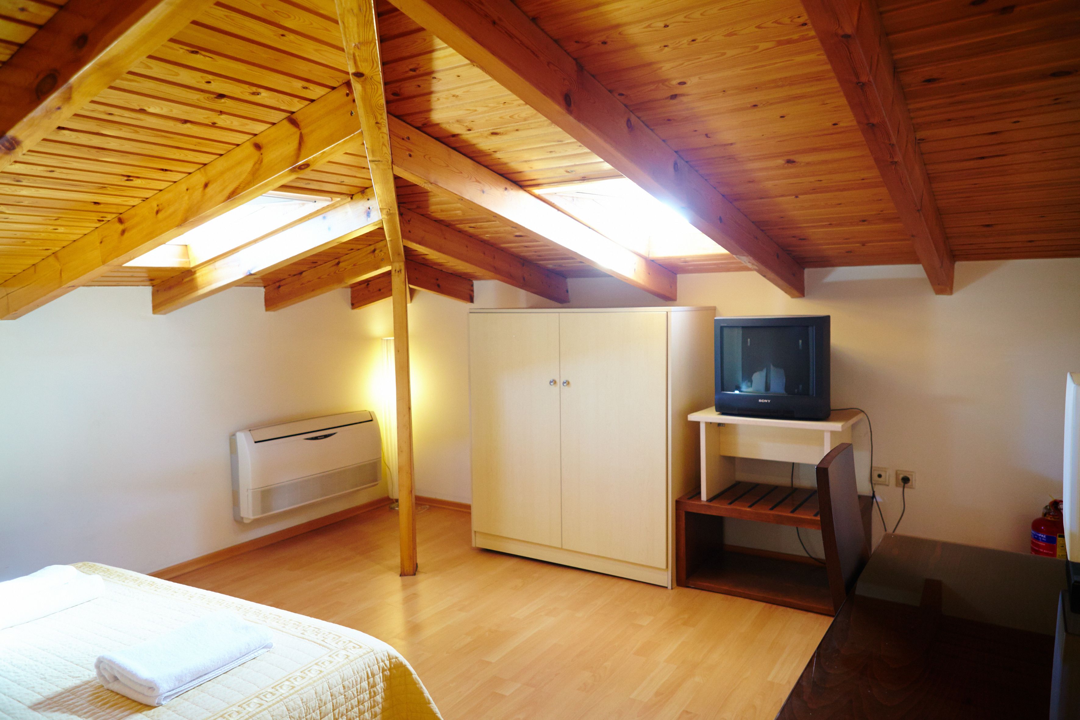 Una habitación con techo inclinado, vigas de madera, una cama con una colcha blanca, un armario, un televisor sobre un soporte y un equipo de aire acondicionado blanco.