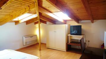Una habitación con techo inclinado, vigas de madera, una cama con una colcha blanca, un armario, un televisor sobre un soporte y un equipo de aire acondicionado blanco.
