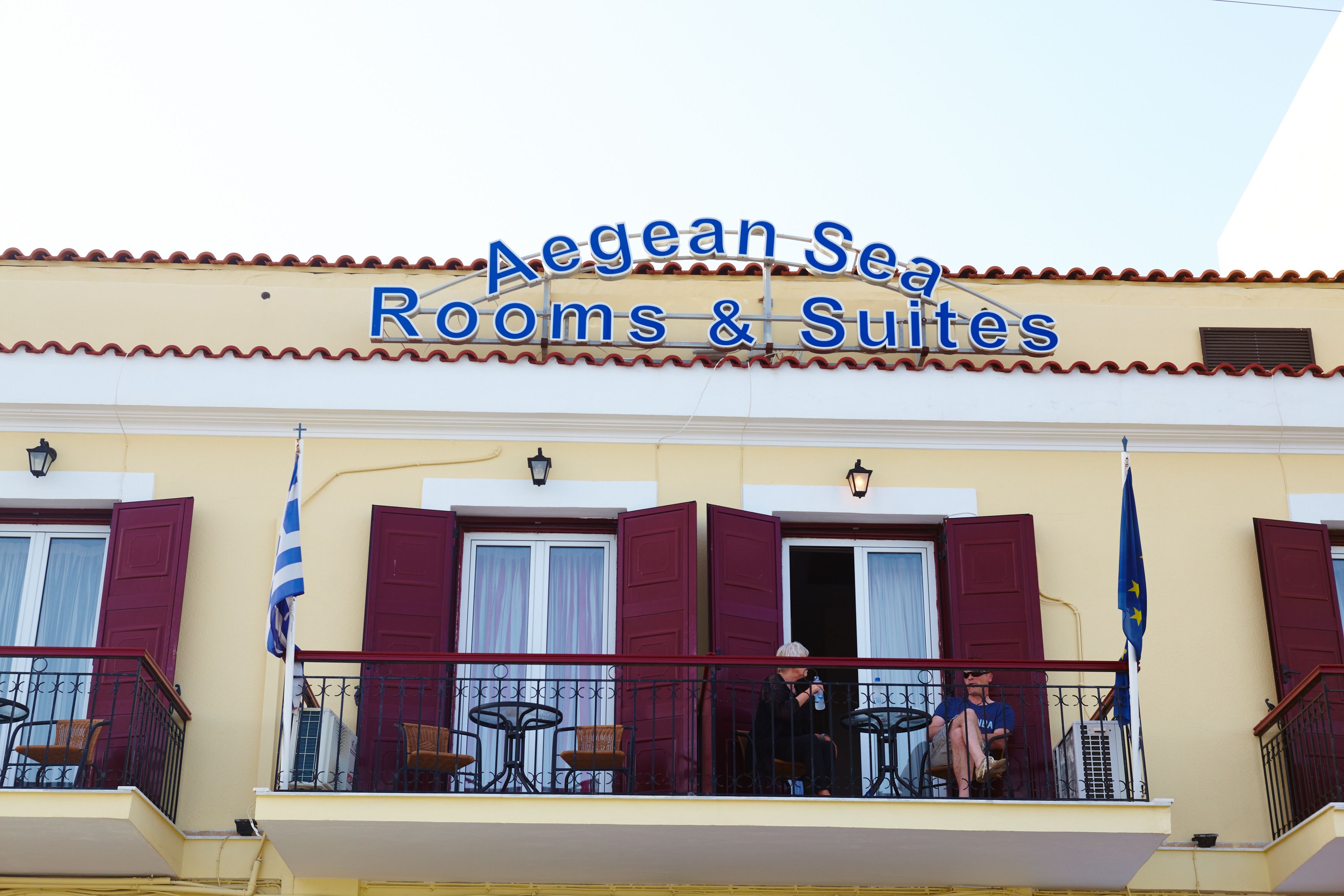 A hotel named "Aegean Sea Rooms & Suites" with a balcony featuring red shutters, outdoor furniture, and a flag.