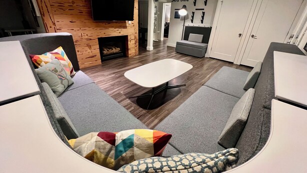 A modern living room with a flat-screen TV mounted on a wooden wall, a fireplace, grey sofas with colorful cushions, and a dining area with a white table and chairs.
