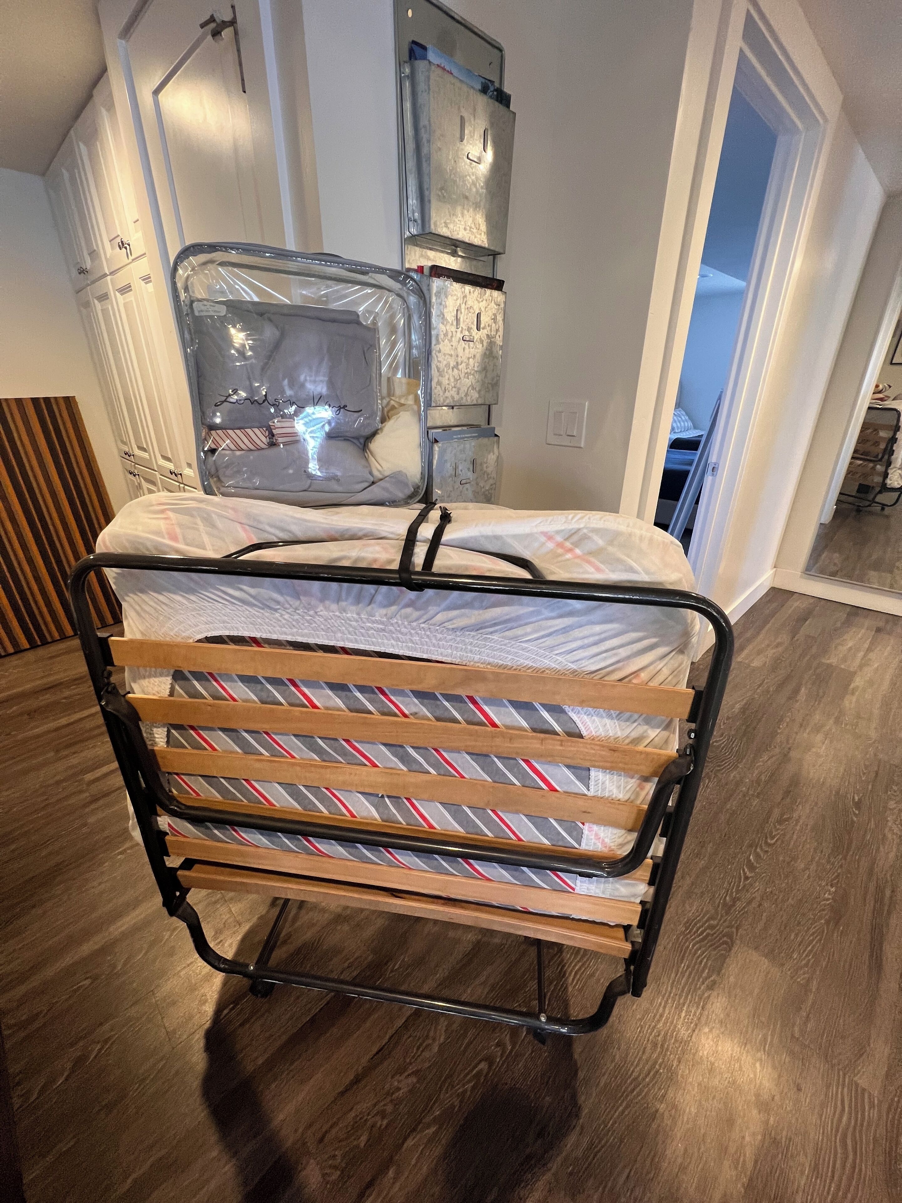 Una cama con estructura metálica y ropa de cama a rayas, suelo de madera y pared blanca con una puerta y un interruptor de luz.