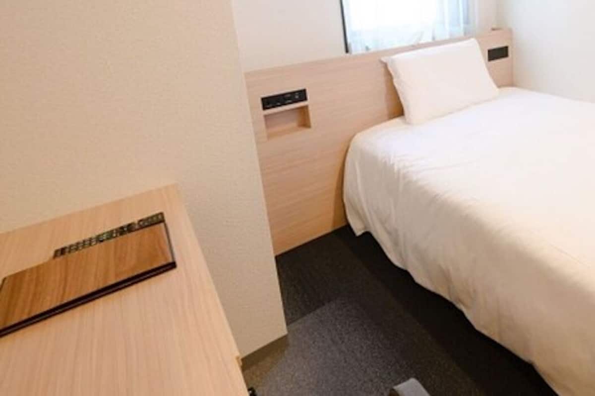 A hotel room with a bed, a small desk, a window with curtains, and a carpeted floor.
