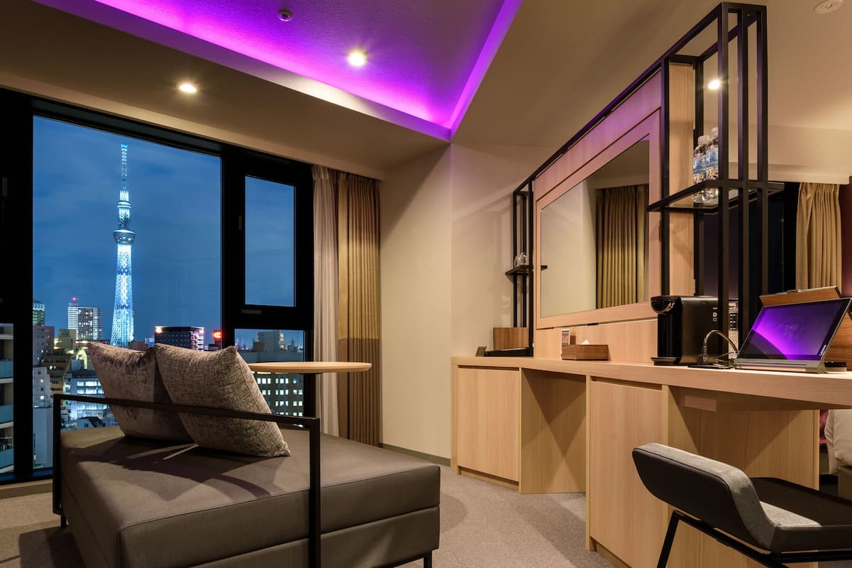 A modern hotel room with a large window offering a city view, a desk with a computer, and a comfortable seating area.