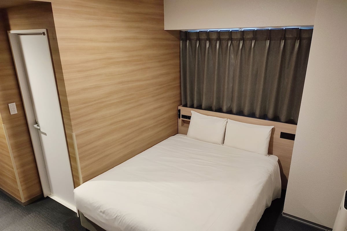 A hotel room with a wooden wall, a bed with white bedding, a window with curtains, and a door.