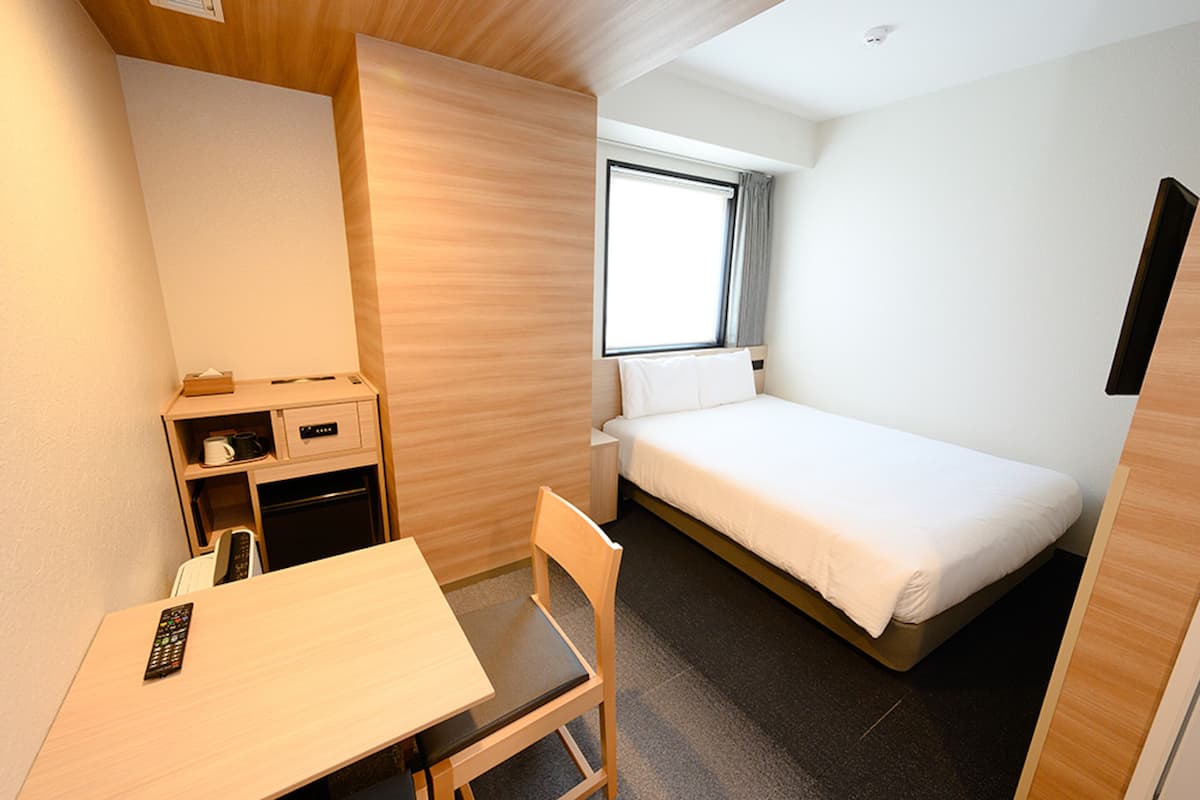 A hotel room with a single bed, a small desk with a chair, a bedside table, and a window with curtains.