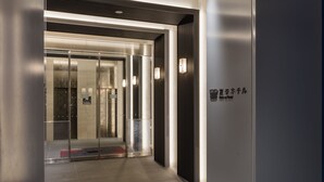 A modern elevator with glass doors and a metallic interior, leading to a lobby area with a light fixture and a sign on the wall. -  Henn na Hotel Premier Tokyo Asakusa Tawaramachi (Tokyo)